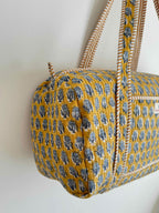 A mustard yellow quilted duffel bag with a blue block-print pattern and cream rope handles, hanging against a plain white wall.
