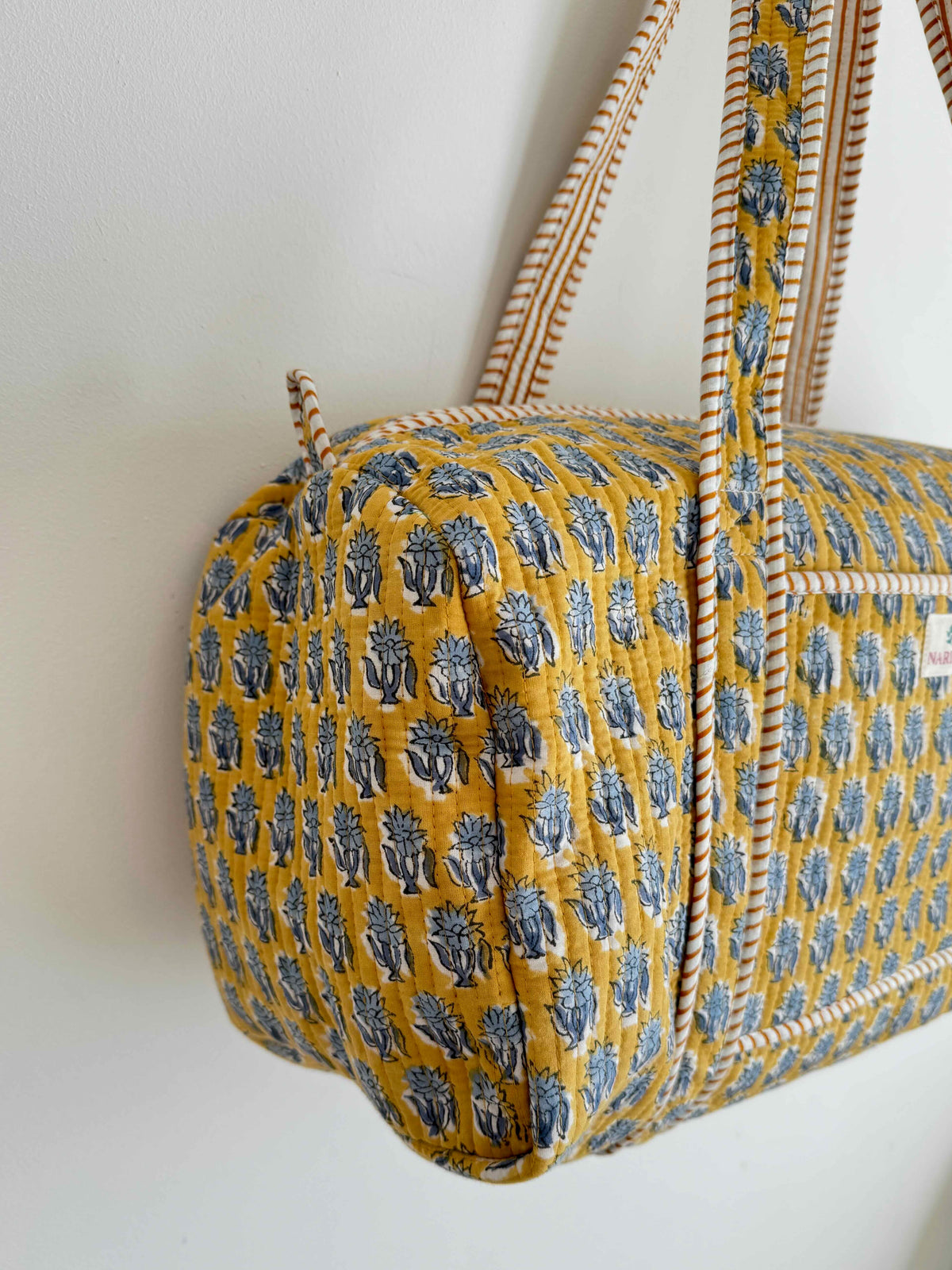 A mustard yellow quilted duffel bag with a blue block-print pattern and cream rope handles, hanging against a plain white wall.